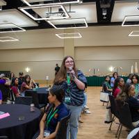 Person standing up in a room of people, all sitting at black tables.